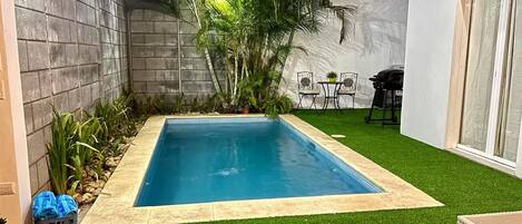 A rectangular pool with a tiled edge, surrounded by artificial grass, with a concrete wall on one side and a white wall on the other.
