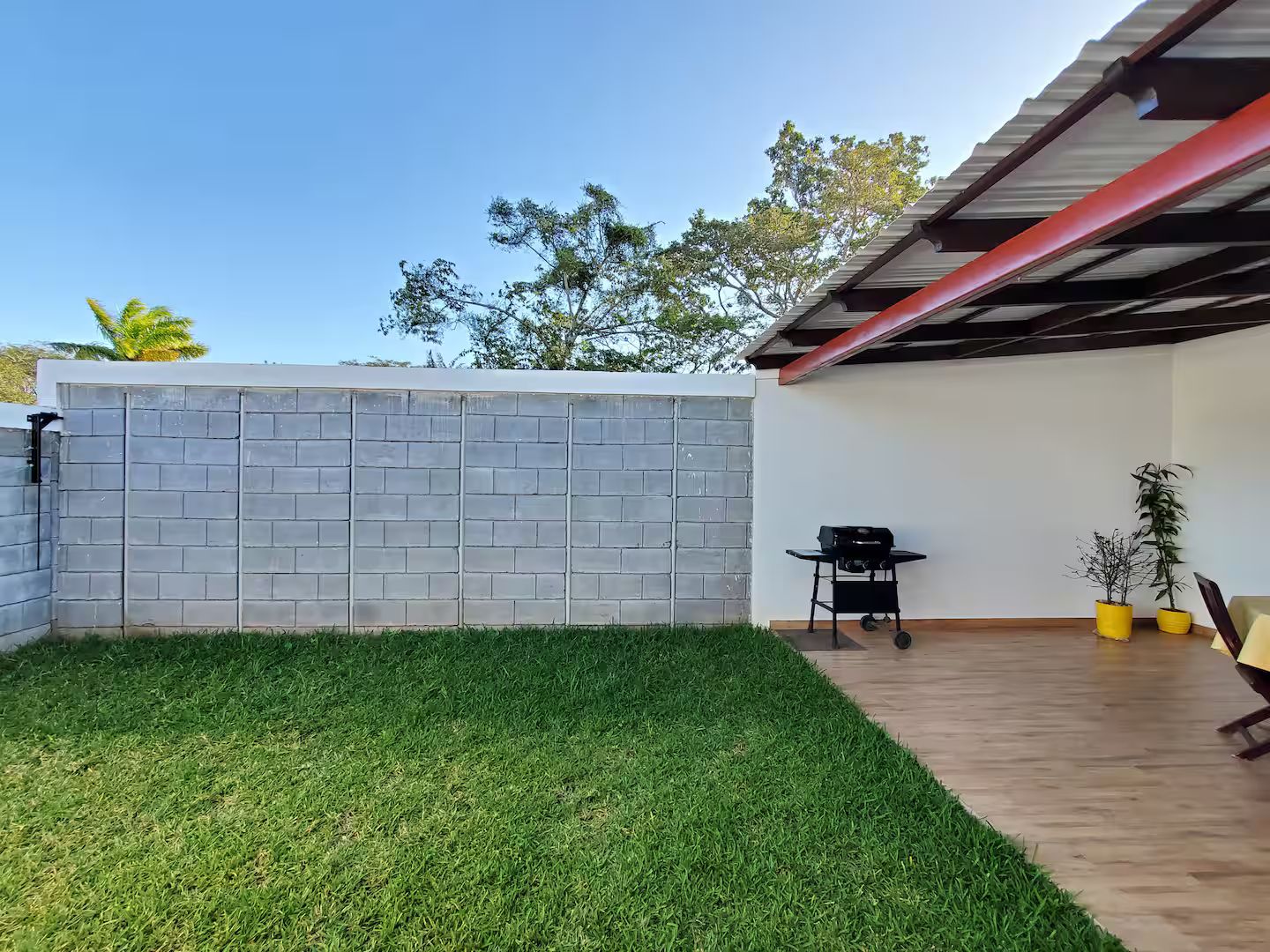 Ein Garten mit Backsteinmauer, Rasenfläche, Grill und Kübelpflanzen.