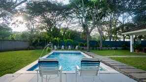 A rectangular swimming pool with two white lounge chairs in the foreground, surrounded by a well-maintained lawn and trees.
