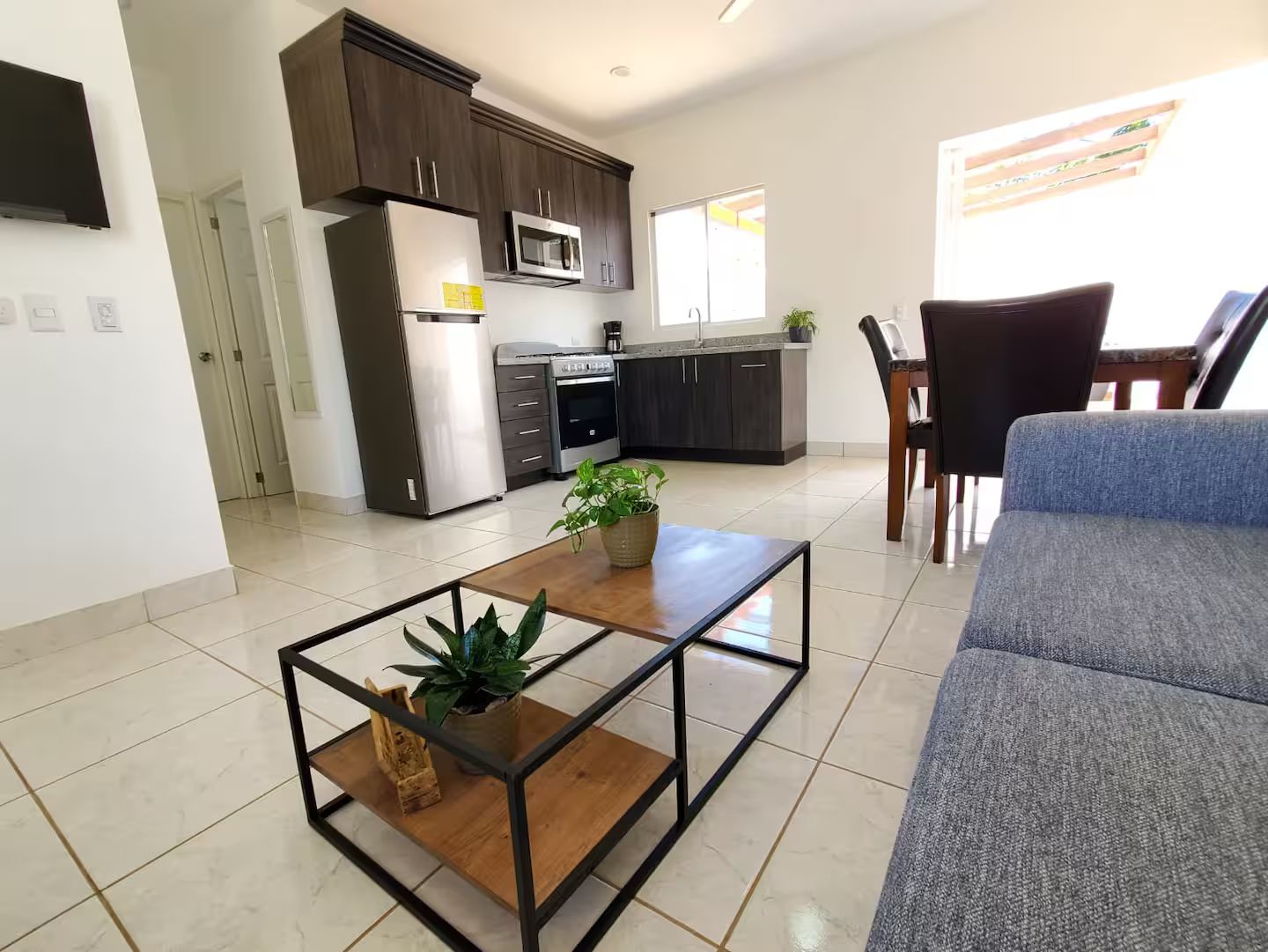 Uma sala de estar moderna com um sofá, uma mesa de centro com plantas e vista para uma cozinha com armários e eletrodomésticos escuros.