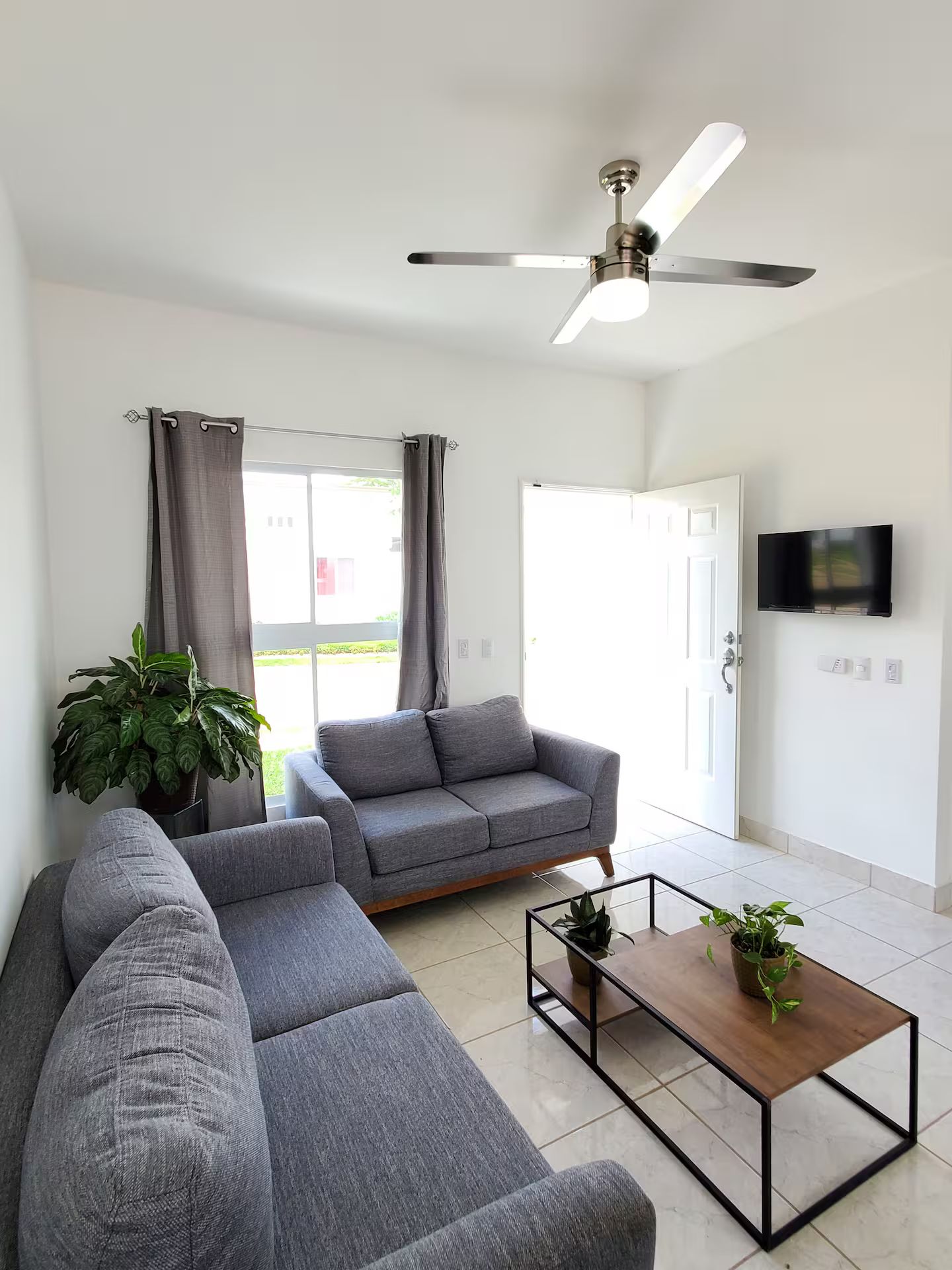 A modern living room with a grey sectional sofa, a coffee table, a ceiling fan, and a TV mounted on the wall.