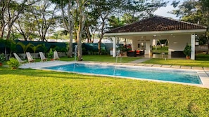 Una piscina con sillones de descanso, un área de estar techada y un césped cuidado.