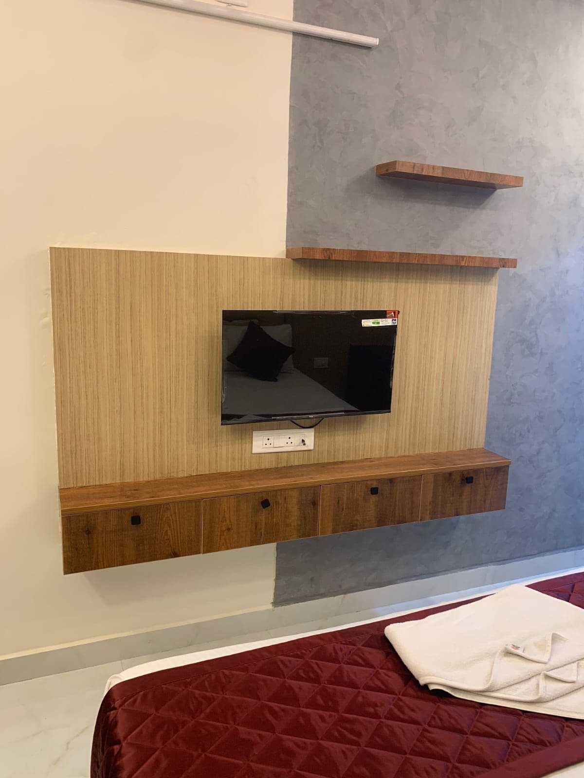 A bedroom with a TV mounted on a wooden panel, a bed with a red quilt, and a grey wall.