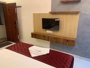 Quarto de hotel com cama, televisão fixada num painel de madeira, porta e pequena estante acima da TV.