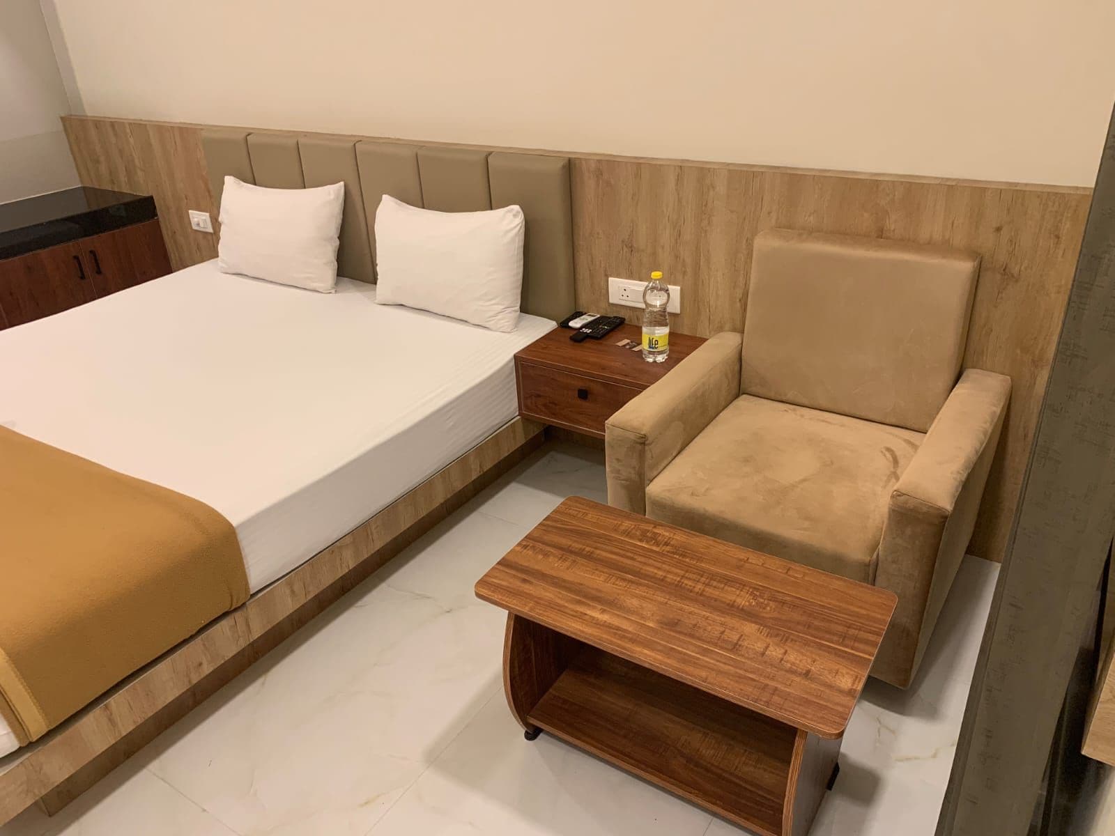 A hotel room with a large bed, a wooden nightstand, a beige armchair, and a small wooden table.