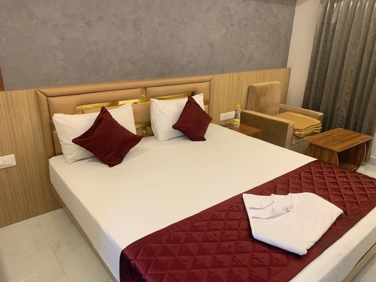 A hotel room with a bed, pillows, a bedside table, a chair, and a window with curtains.