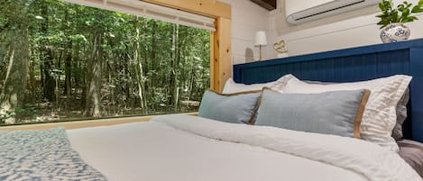 A bedroom with a large bed, a view of a forest through a window, and an air conditioning unit on the wall.