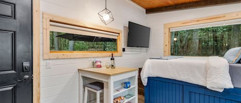 A bedroom with a bed, a desk with a chair, a TV, and a window with a view of trees.