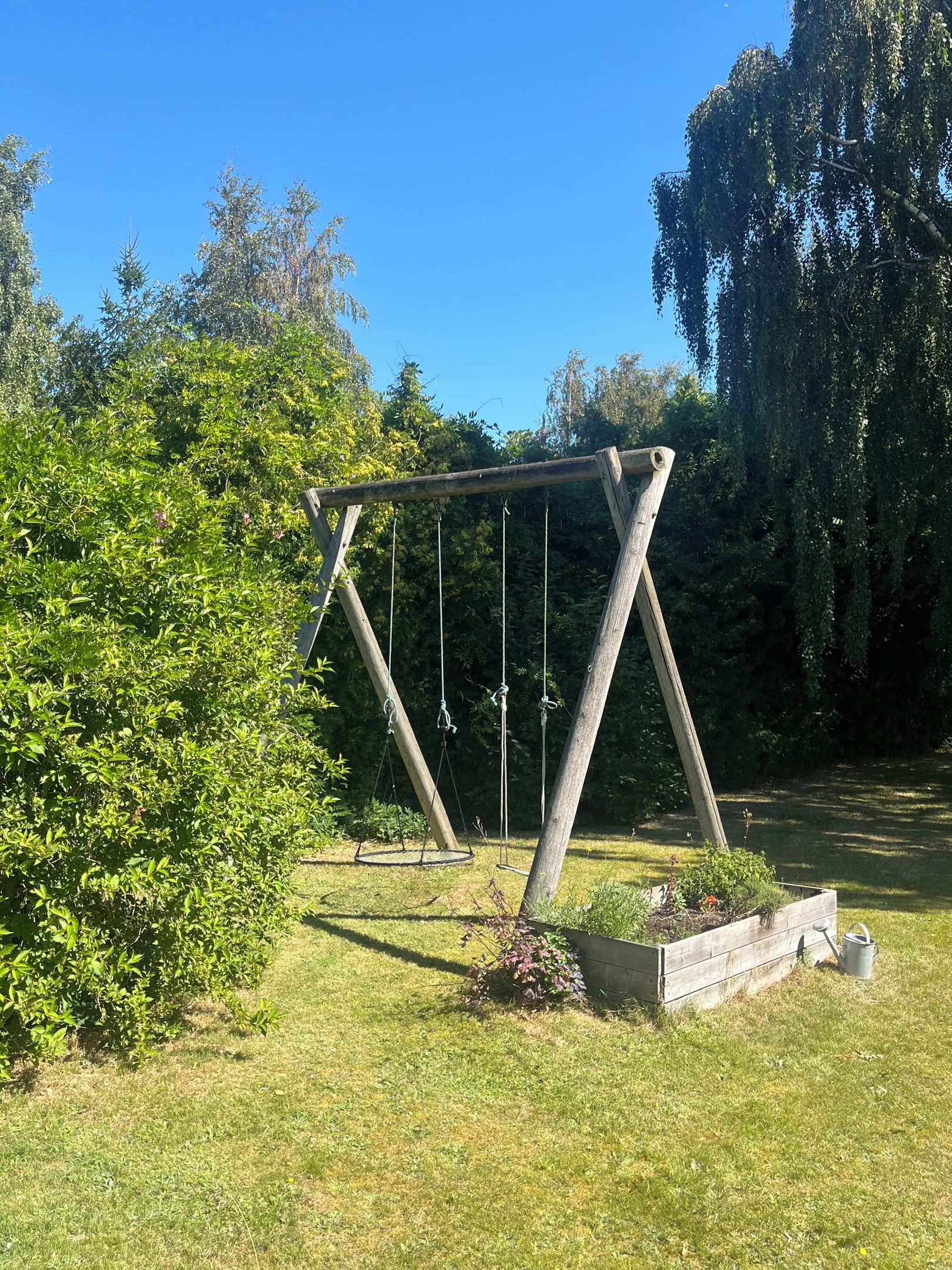 A wooden swing set in a garden with grass and trees.