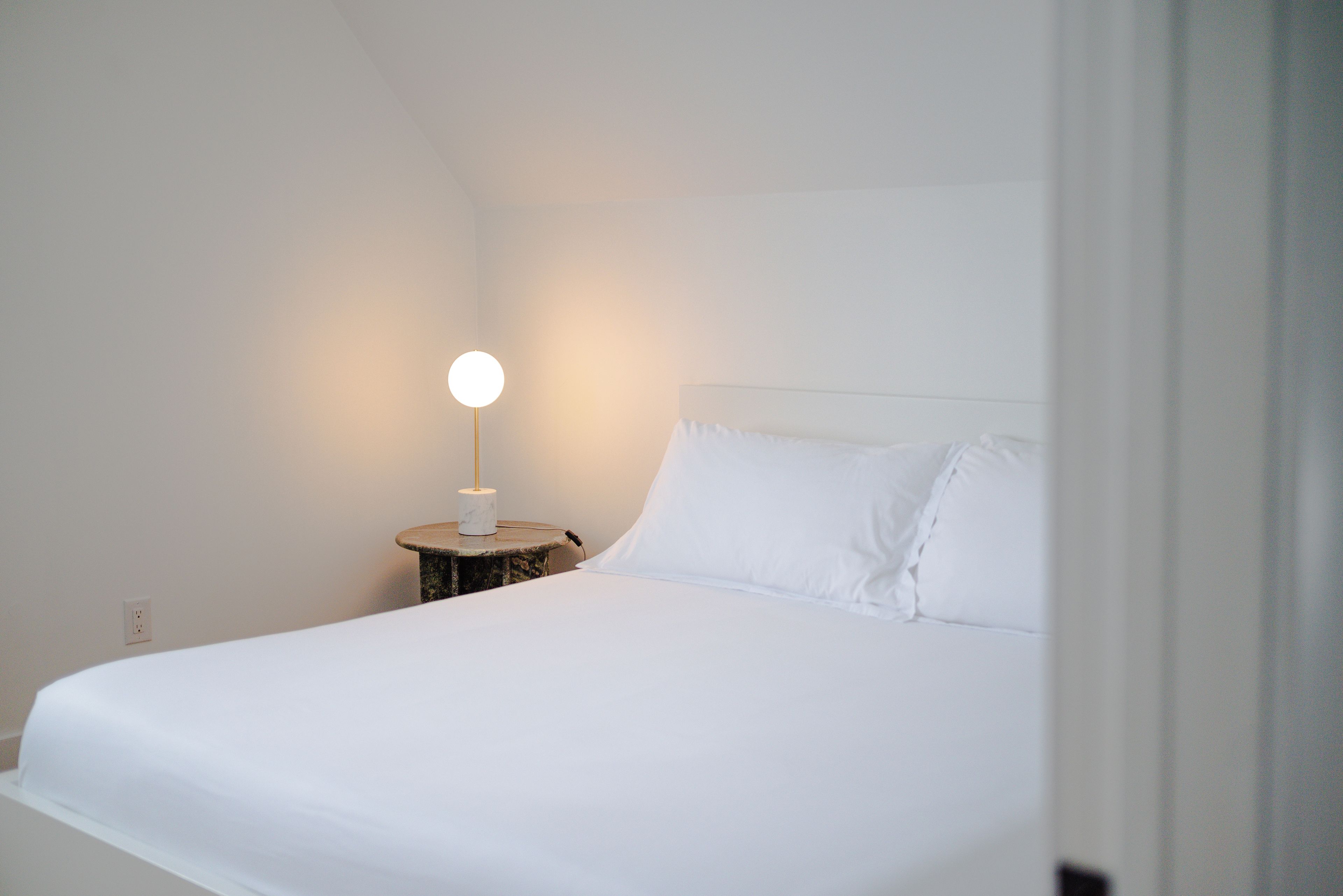 A single bed with white bedding and pillows, a bedside table with a lamp, and a power outlet on the wall.