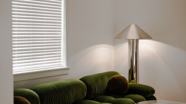 Ein Zimmer mit einer Eckcouch, einer Lampe und einem Fenster mit Jalousien.