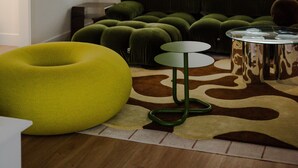 Ein Zimmer mit gewölbter Decke, einem großen Fenster mit Jalousien, einem grünen Sofa, einem gelben Sitzsack und einem kleinen runden Tisch mit Glasplatte.