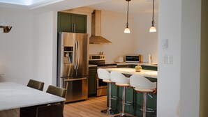 Eine moderne Küche mit einem Edelstahl-Kühlschrank, einer Mikrowelle und einem Einbauofen. Zwei Barhocker und ein Esstisch mit Stühlen sind vorhanden.
