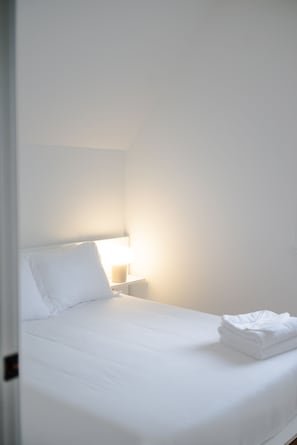 Un lit simple avec du linge de lit blanc, dans une pièce aux murs blancs et dotée d’une table de chevet.