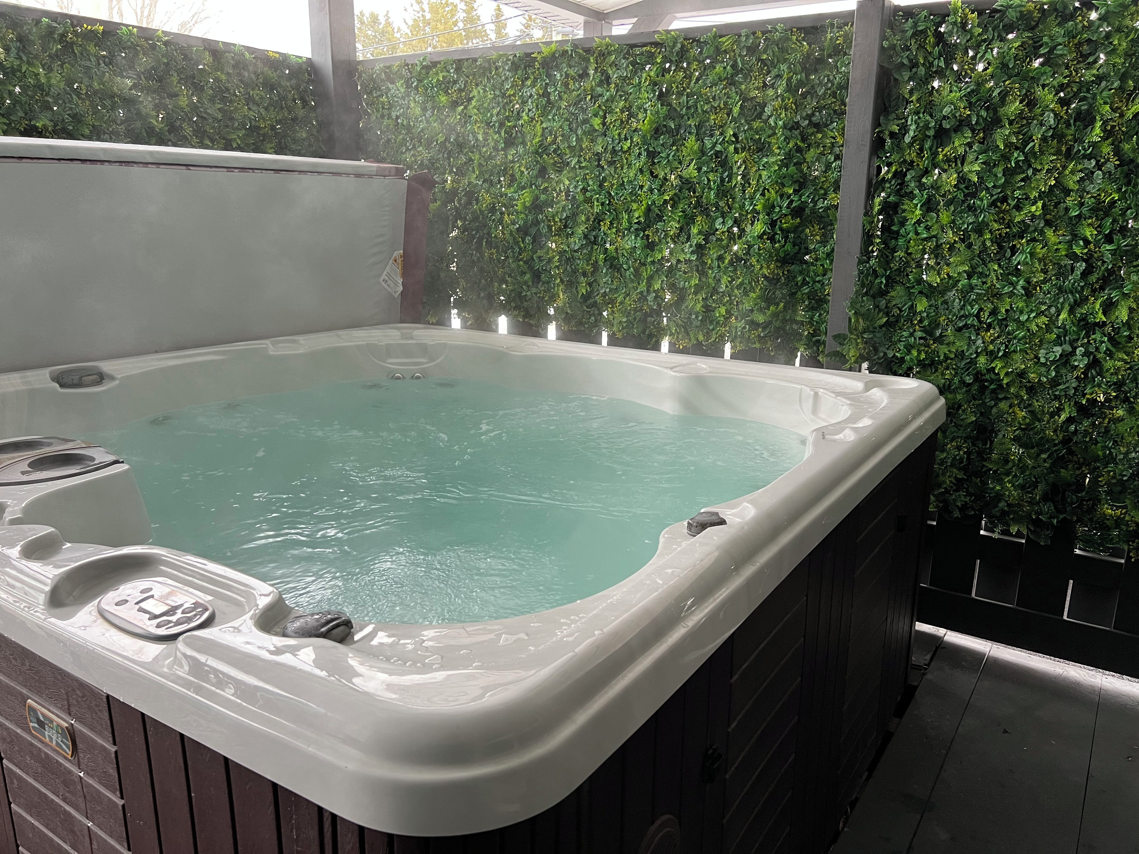 A hot tub with a wooden exterior, surrounded by a wall of green foliage.