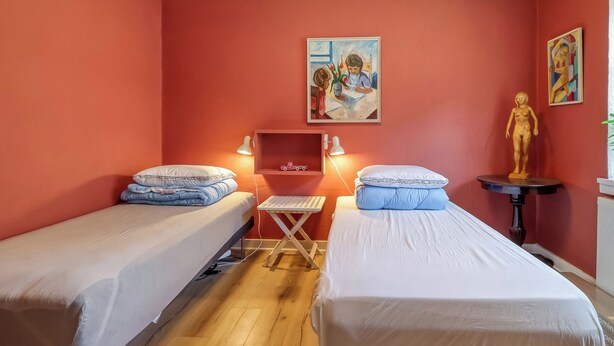 Une chambre avec deux lits simples, une petite table, une étagère fixée au mur avec un jouet, et un tableau.