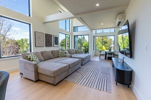 Ein modernes Wohnzimmer mit einer Eckcouch, einem Flachbildfernseher und einem großen Fenster mit Blick auf Bäume.