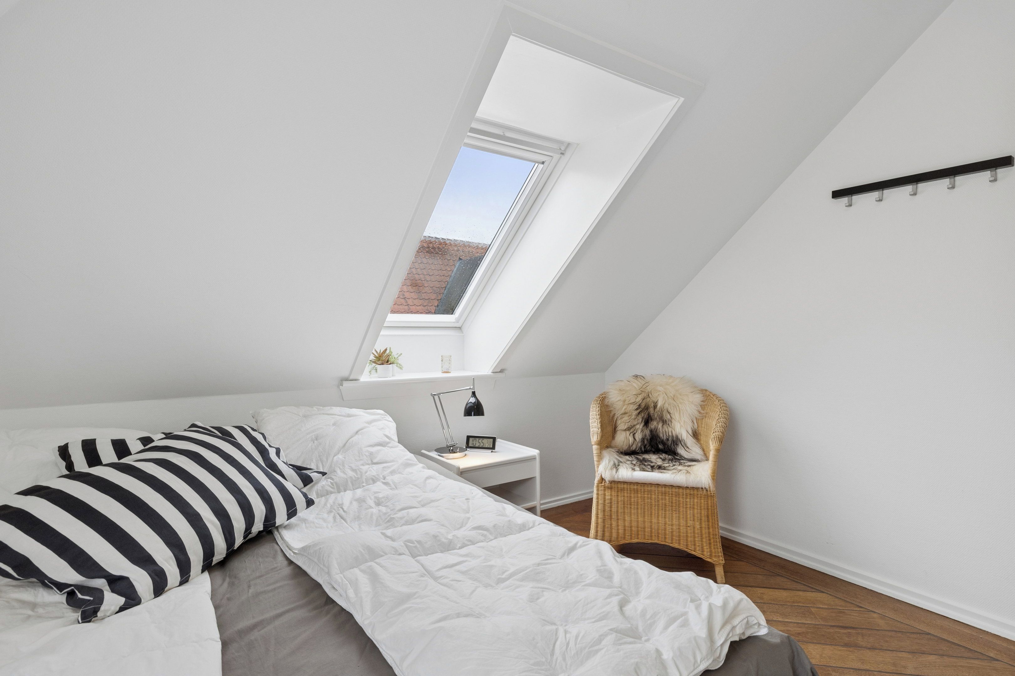 A bedroom with a sloped ceiling, a bed with white bedding, a nightstand with a lamp, a chair, and a window with a view of a red-roofed building.