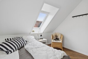 A bedroom with a sloped ceiling, a bed with white bedding, a nightstand with a lamp, a chair, and a window with a view of a red-roofed building. - Lovely apartment in the heart of Skagen town (Skagen)