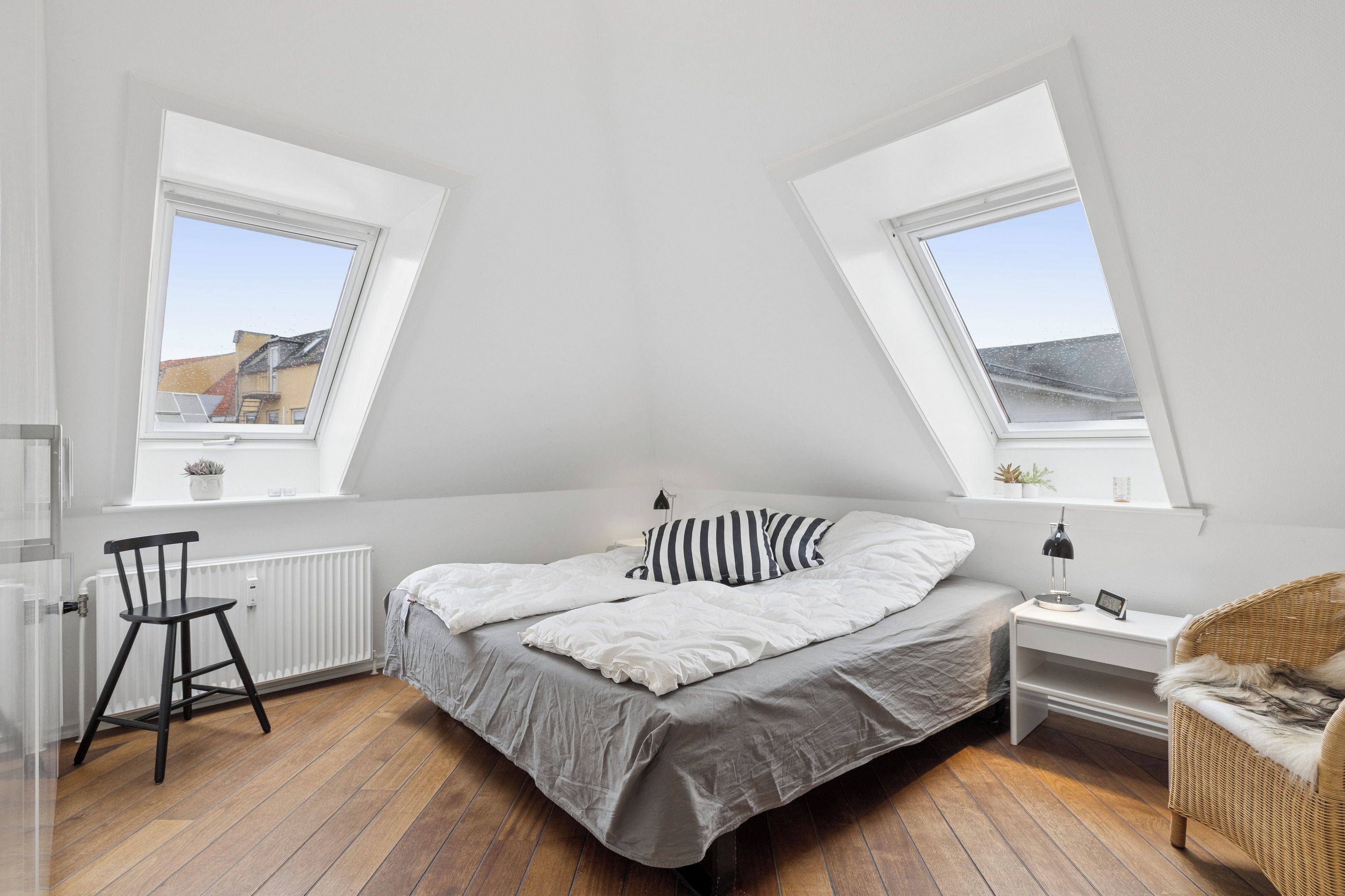 A bedroom with a bed, a chair, a nightstand, a lamp, and two windows.