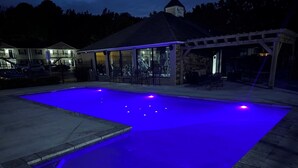 En swimmingpool med blått ljus, en täckt uteplats och en byggnad med ett garage i bakgrunden.