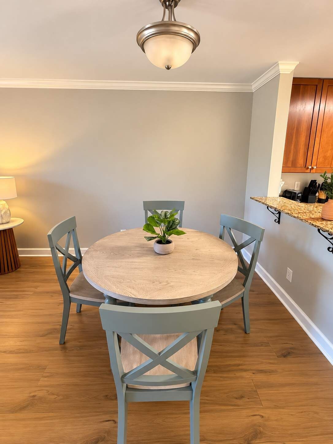 A dining area with a round table, four chairs, a ceiling light, a small plant, and a kitchen area with wooden cabinets.