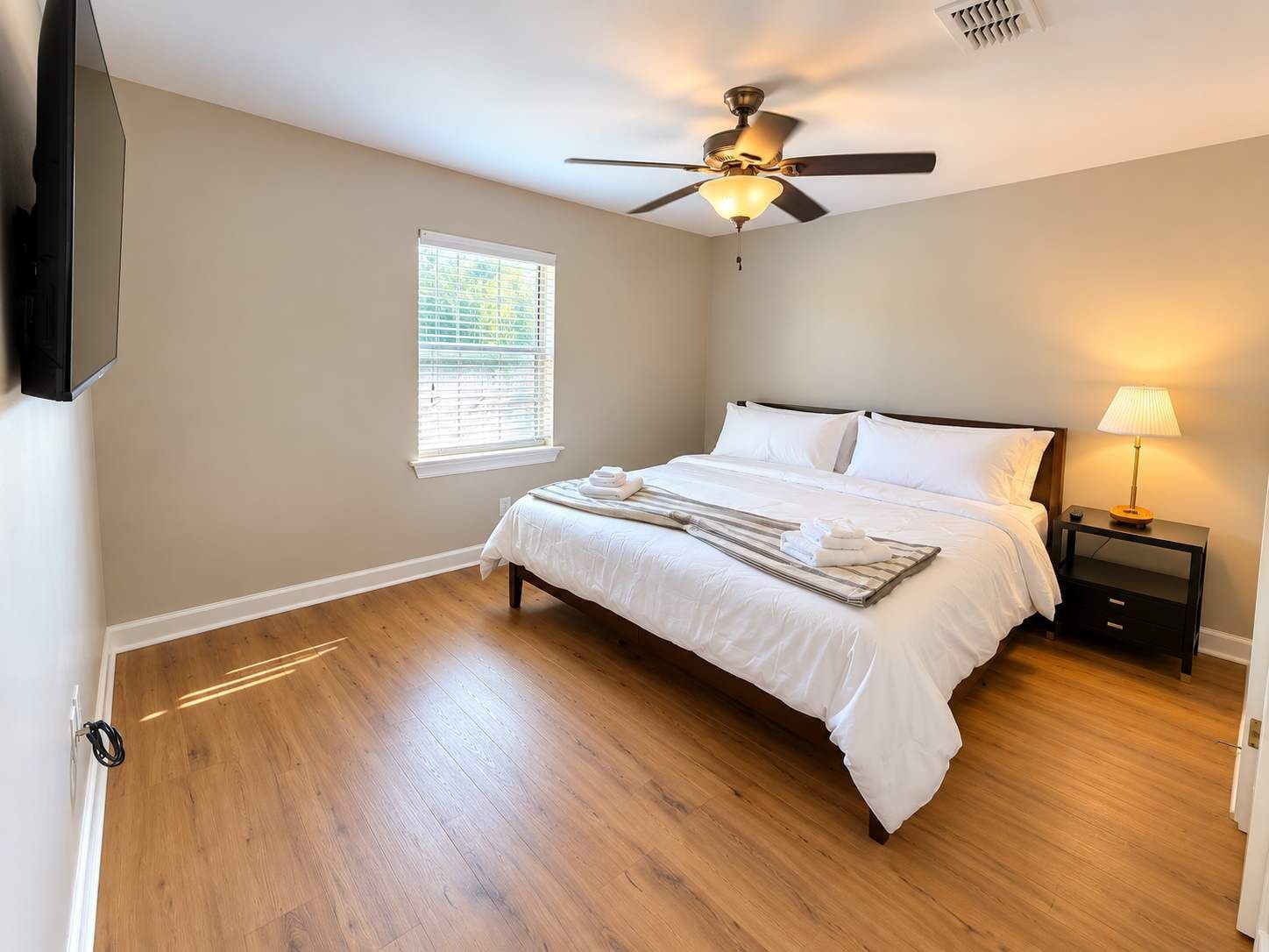 A bedroom with a bed, a ceiling fan, a nightstand with a lamp, and a window with blinds.