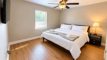 A bedroom with a bed, a ceiling fan, a nightstand with a lamp, and a window with blinds.