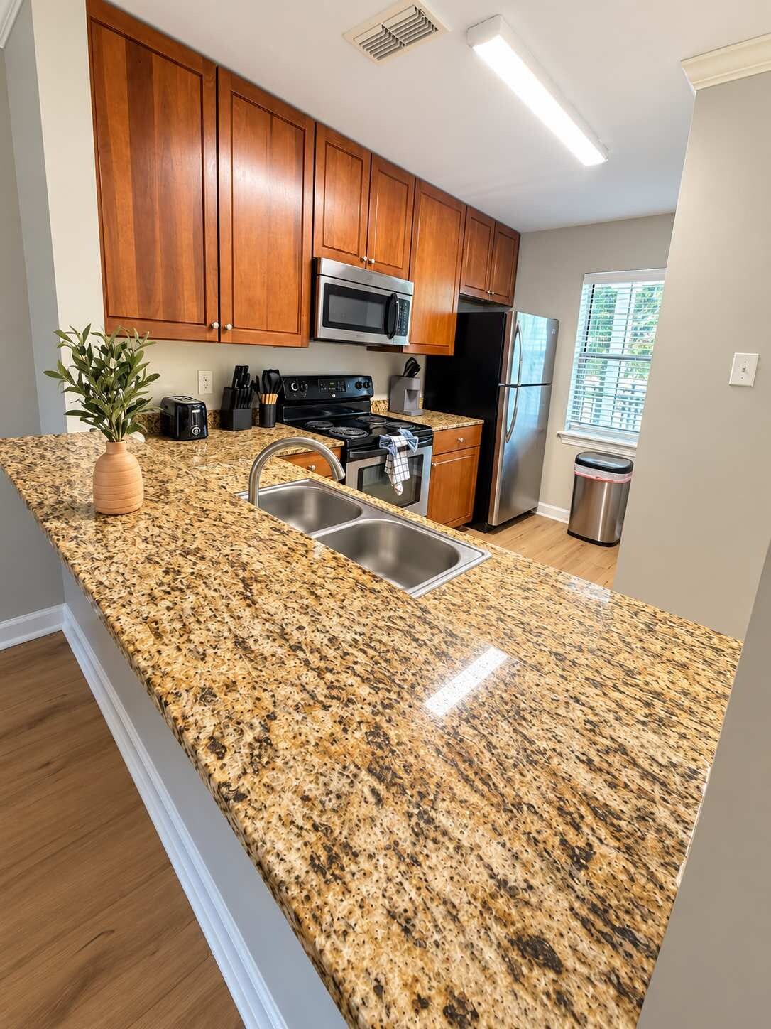 Una cocina con encimeras de granito, armarios de madera, electrodomésticos de acero inoxidable y una ventana con persianas.