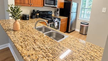 Una cocina con encimeras de granito, armarios de madera, electrodomésticos de acero inoxidable y una ventana con persianas.