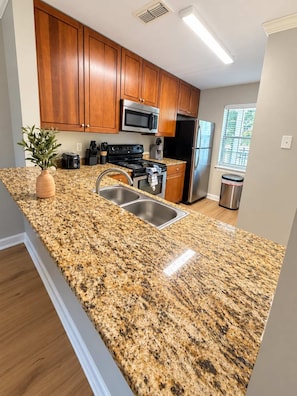 A kitchen with granite countertops, wooden cabinets, stainless steel appliances, and a window with blinds.