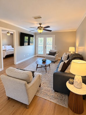 A living room with a sofa, armchair, coffee table, and a lamp. There is a ceiling fan and a TV mounted on the wall.