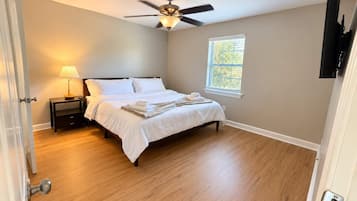 Un dormitorio con cama, mesita de noche, lámpara, ventilador de techo, ventana y televisor montado en la pared.
