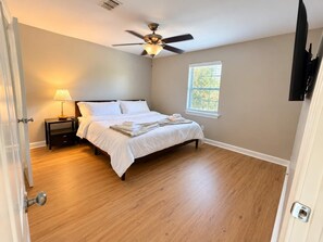 A bedroom with a bed, a nightstand, a lamp, a ceiling fan, a window, and a TV mounted on the wall.