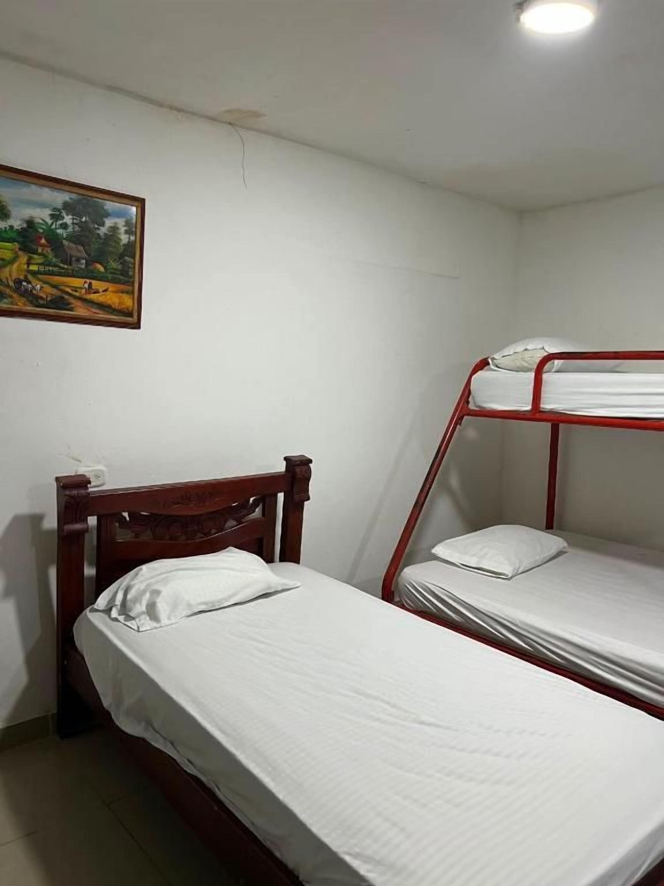 A room with two beds, one with a wooden frame and one with a metal frame, a painting on the wall, and a ceiling light.