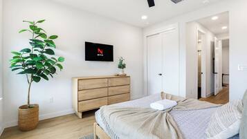 Un dormitorio con cama, televisor montado en la pared, cómoda de madera, planta en maceta y una puerta que da a otra habitación.