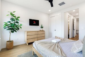 A bedroom with a bed, a TV mounted on the wall, a wooden dresser, a potted plant, and a door leading to another room. - Luxury Condo With Pool-view Balcony (New Braunfels)