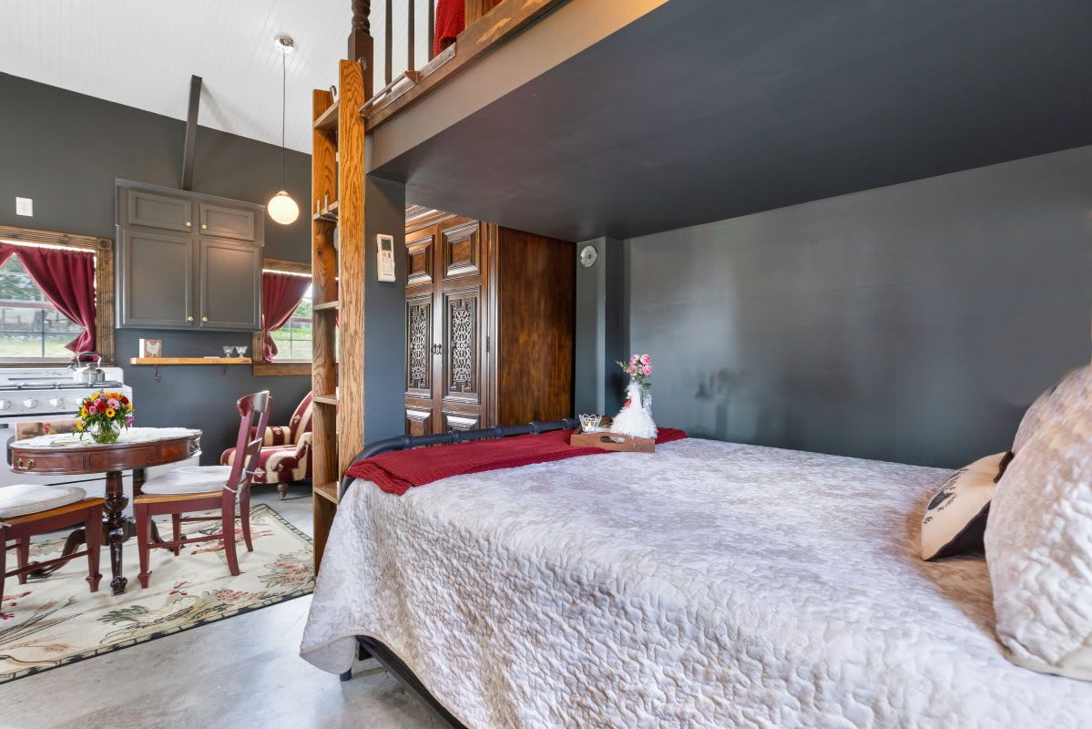 Une chambre avec un grand lit, une chaise en bois, une petite table avec un vase, et une vue sur la cuisine.