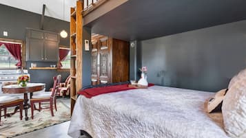 Une chambre avec un grand lit, une chaise en bois, une petite table avec un vase, et une vue sur la cuisine.