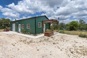 A green building with a garage, a wooden pergola, and a gravel driveway. - The Stargazer Cabin - A Boutique Hill Country Retreat (Fredericksburg)