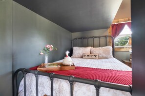 A bedroom with a bed, a nightstand, a vase with flowers, and a window with curtains. - The Stargazer Cabin - A Boutique Hill Country Retreat (Fredericksburg)