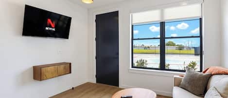 Una sala de estar moderna con un televisor montado, un sofá, una mesa de centro y un amplio ventanal con vistas a un campo deportivo.