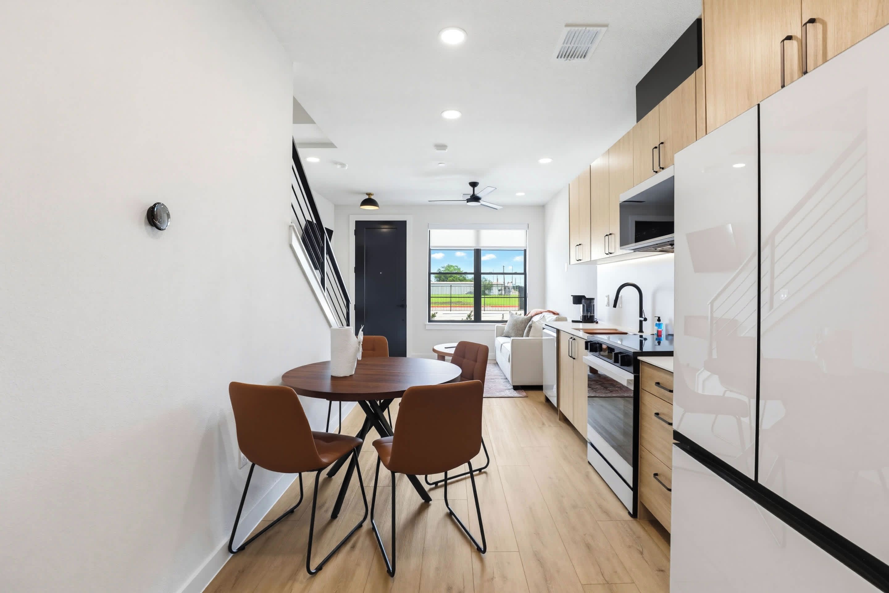Una cocina moderna con armarios blancos, un comedor con sillas marrones y vistas por la ventana.
