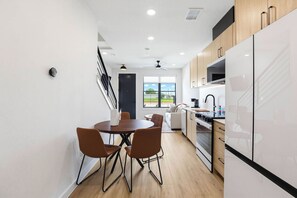 A modern kitchen with white cabinets, a dining area with brown chairs, and a view through the window. - Prism at Dovetail - 1102 (New Braunfels)