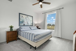 Une chambre à coucher avec un lit, une table de chevet, un ventilateur de plafond, une fenêtre avec des rideaux et un tableau au mur.