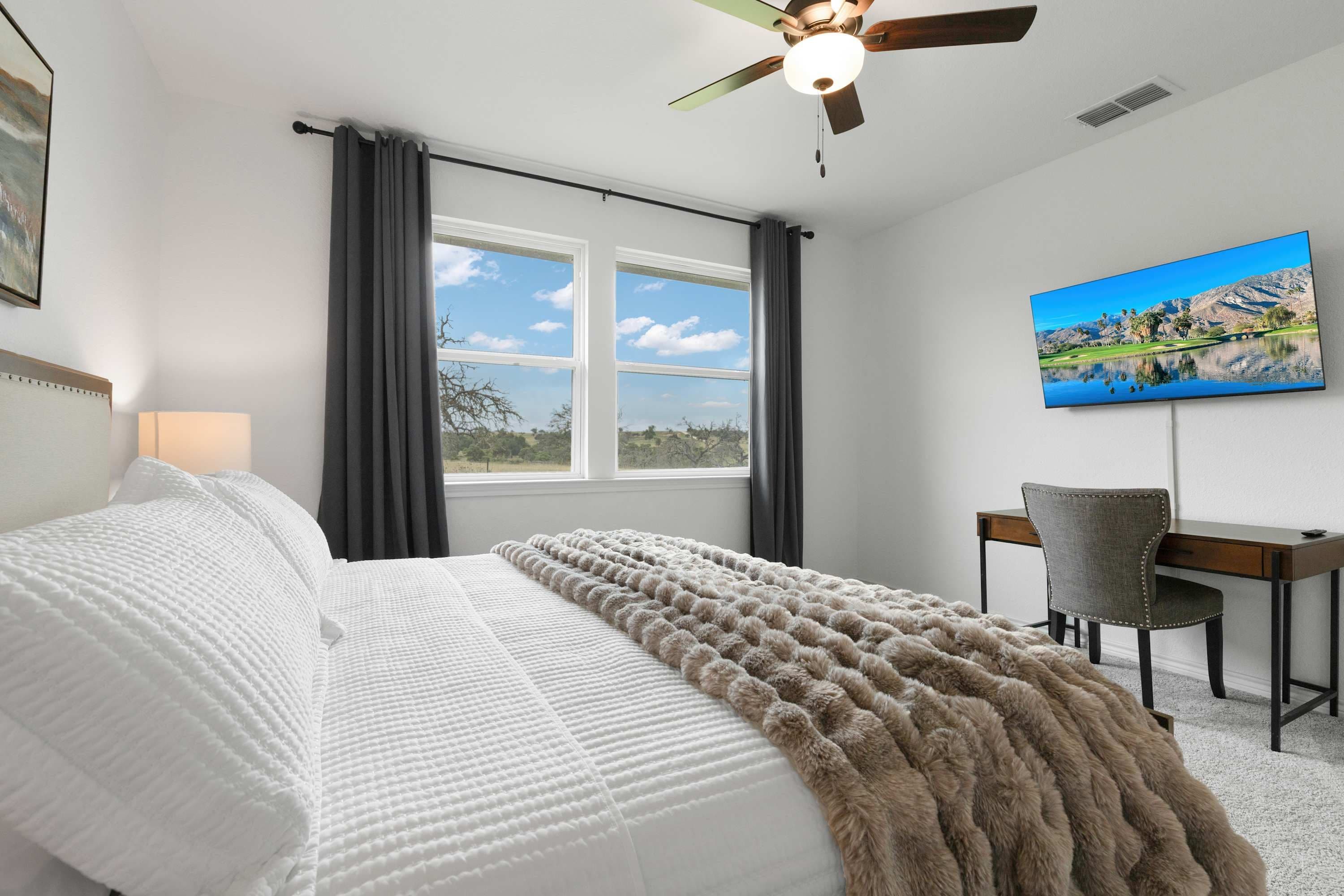 A bedroom with a bed, a desk, a chair, a ceiling fan, a window with curtains, and a painting on the wall.