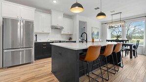 A modern kitchen with a central island, stainless steel appliances, and a dining area with bar stools. - Birdsong Under the Stars Hot Tub, Fire Pit & Hill Country Views (Fredericksburg)