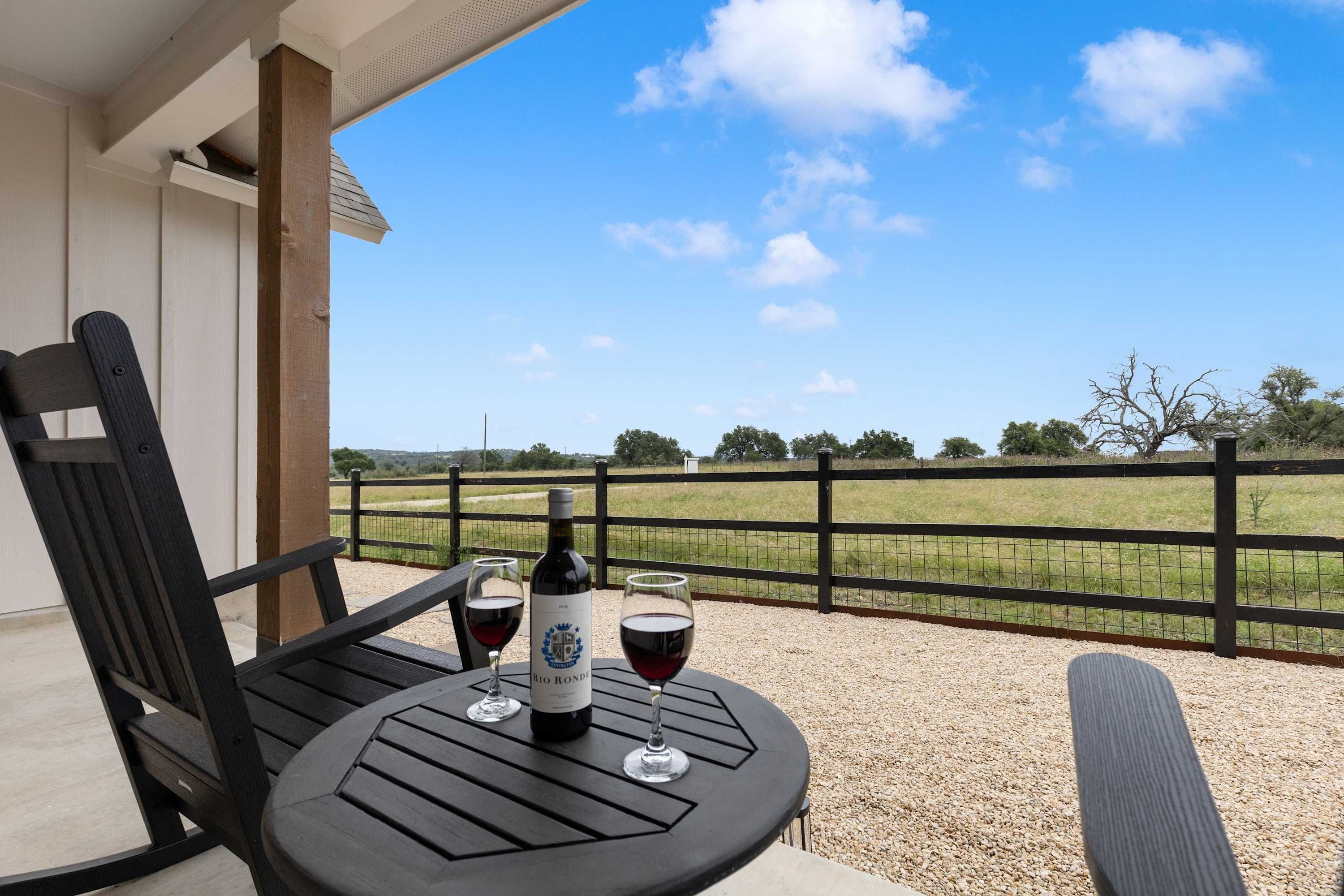Une table avec deux verres à vin et une bouteille, placée sur un balcon donnant sur un champ clôturé.