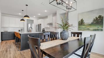 A modern kitchen with white cabinets, a central island, pendant lighting, and a dining area with a wooden table and chairs.