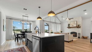 A modern kitchen with a central island, dining area, and a fireplace. - Birdsong Under the Stars Hot Tub, Fire Pit & Hill Country Views (Fredericksburg)